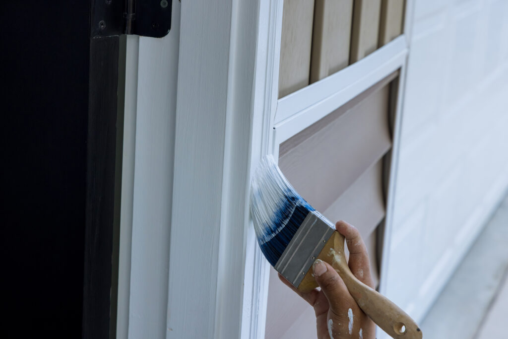 Close-up of a painter's hand holding a flat brush applying white paint to wooden trim beside a door frame.